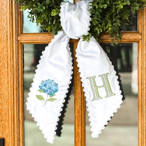 May include: White fabric door hanger with a scalloped edge, featuring a blue hydrangea flower and the letter "H" in green embroidery. The hanger is displayed on a wooden door with a green wreath.