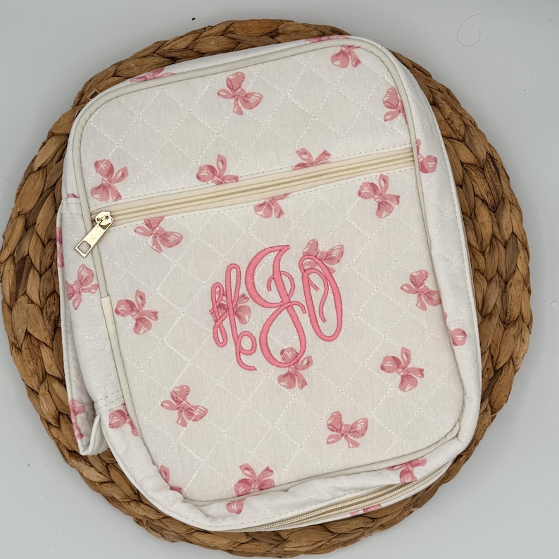 May include: A quilted, off-white backpack with a repeating pattern of pink bows. The backpack features a gold zipper and a pink monogram. The bag is set on a woven, circular placemat.