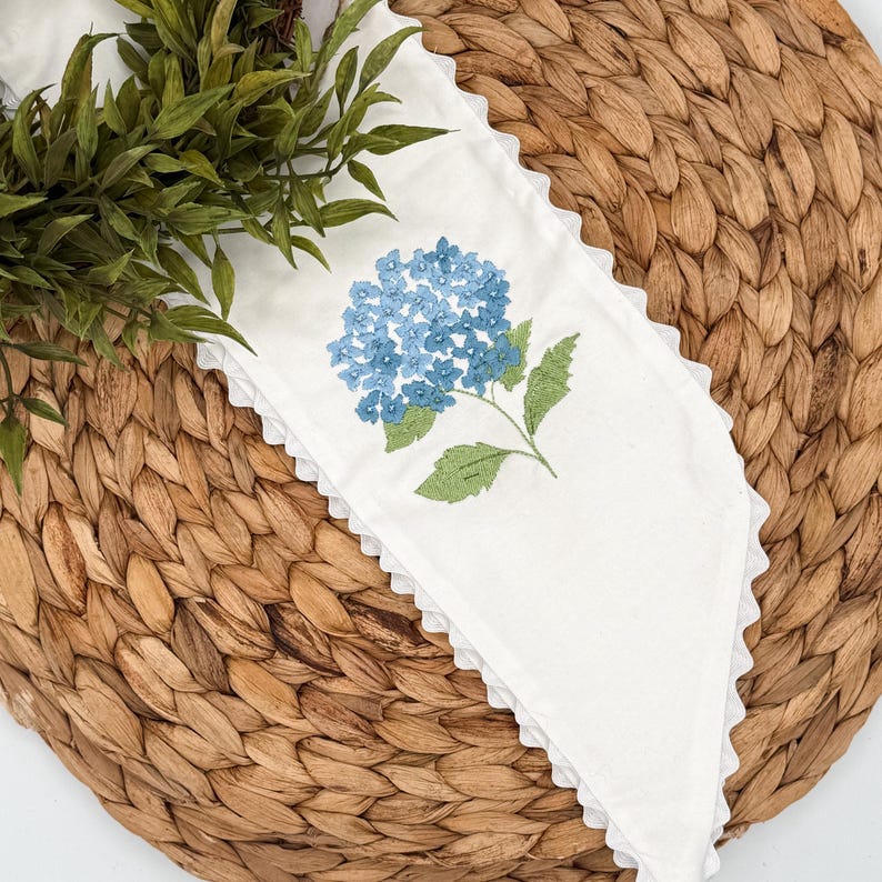 May include: White linen napkin with a blue hydrangea embroidery and a scalloped edge. The napkin is placed on a woven, brown placemat, with green foliage in the background. The floral design adds a touch of elegance.
