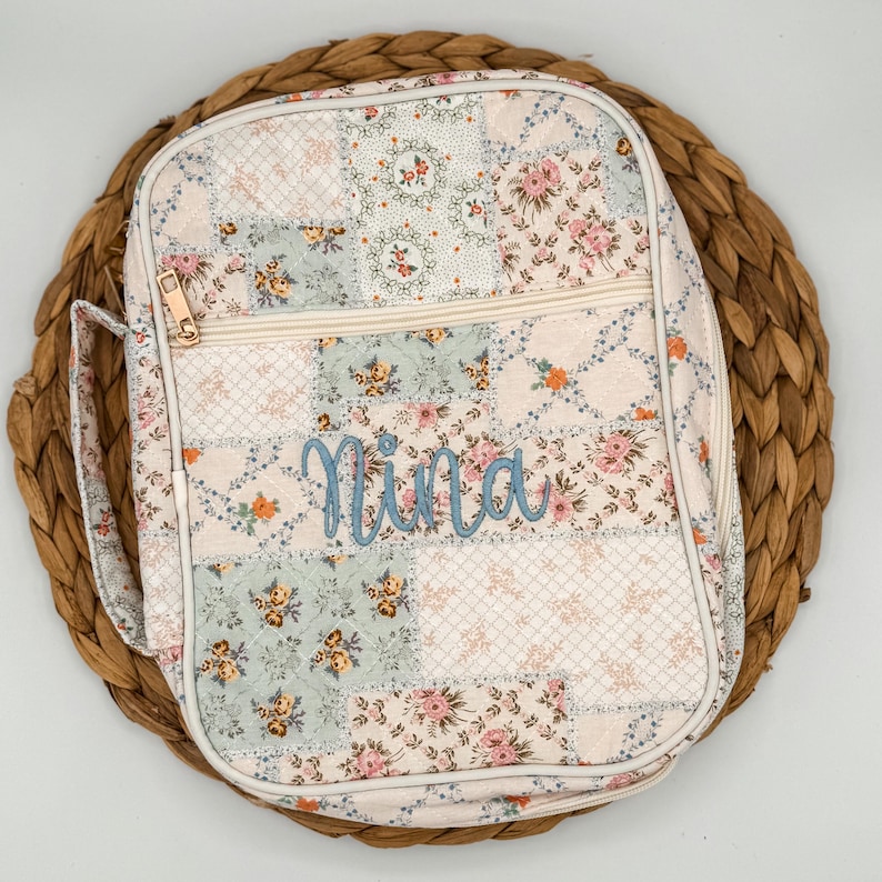 May include: A floral patchwork lunch bag with a zipper closure and a handle. The bag features a variety of pastel floral patterns and the name "Nina" embroidered in light blue script.