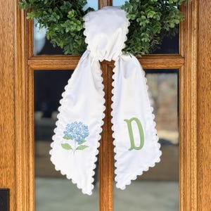 May include: White door sash with a scalloped edge, adorned with a blue hydrangea and a green letter "D". The sash is tied to a green wreath, creating a decorative accent for a door or entryway.