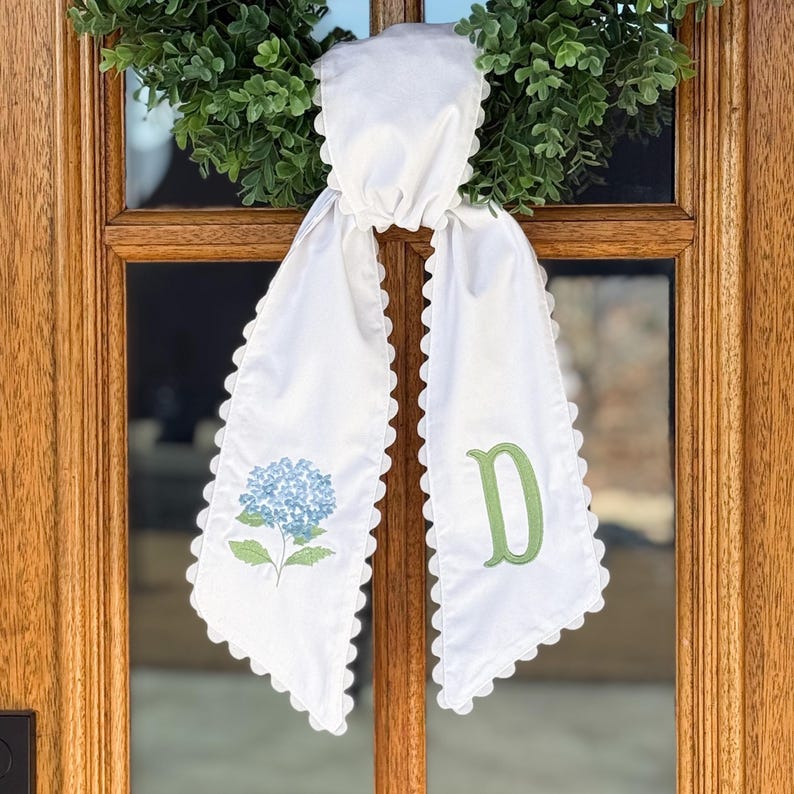 May include: White door sash with a scalloped edge, adorned with a blue hydrangea and a green letter "D". The sash is tied to a green wreath, creating a decorative accent for a door or entryway.