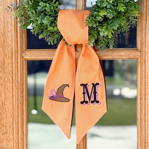 May include: Orange polka dot fabric door hanger with a witch hat and the letter "M" embroidered on it. The door hanger is hanging on a wooden door with a green wreath.