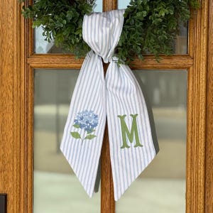 May include: A white and blue striped door sash with a blue hydrangea flower and green letter "M" design. The sash is hanging on a wooden door with a green wreath.