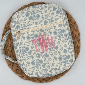 May include: A floral print bag with a handle and zipper closure. The bag features a light cream background with blue flower patterns and a pink monogram. The bag is sitting on a woven mat.