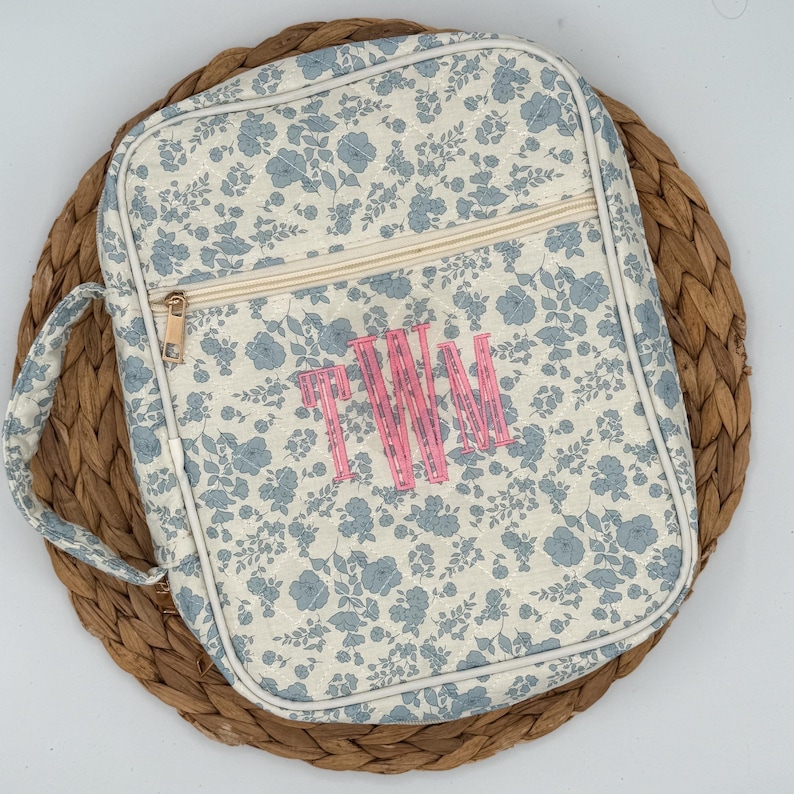 May include: A floral print bag with a handle and zipper closure. The bag features a light cream background with blue flower patterns and a pink monogram. The bag is sitting on a woven mat.