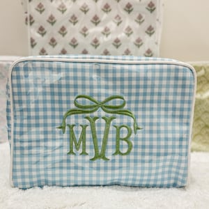 May include: A blue and white gingham cosmetic bag with a white trim. The bag features a green embroidered bow and the initials "MVB" in green. Other bags with floral patterns are visible in the background.