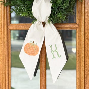 May include: A cream-colored fabric ribbon tied in a knot, featuring a printed orange pumpkin with a green bow on the left tail and a green letter K on the right tail, positioned over a green wreath hanging on a wooden door frame.