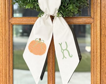 May include: A cream-colored fabric ribbon tied in a knot, featuring a printed orange pumpkin with a green bow on the left tail and a green letter K on the right tail, positioned over a green wreath hanging on a wooden door frame.