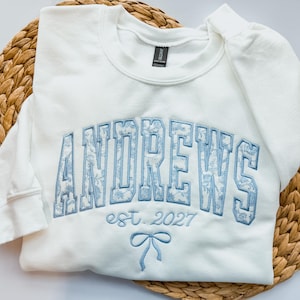 May include: A white crewneck sweatshirt featuring the word ANDREWS in large, light blue block letters with a floral pattern fill. Below the text, the words est. 2027 are embroidered in light blue, followed by a small light blue bow graphic.