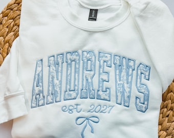 May include: A white crewneck sweatshirt featuring the word ANDREWS in large, light blue block letters with a floral pattern fill. Below the text, the words est. 2027 are embroidered in light blue, followed by a small light blue bow icon.