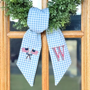 May include: A light blue and white gingham fabric ribbon tied in a bow. The left tail features an embroidered design of two crossed American flags with a dark blue bow, and the right tail features a large embroidered red and blue letter W.