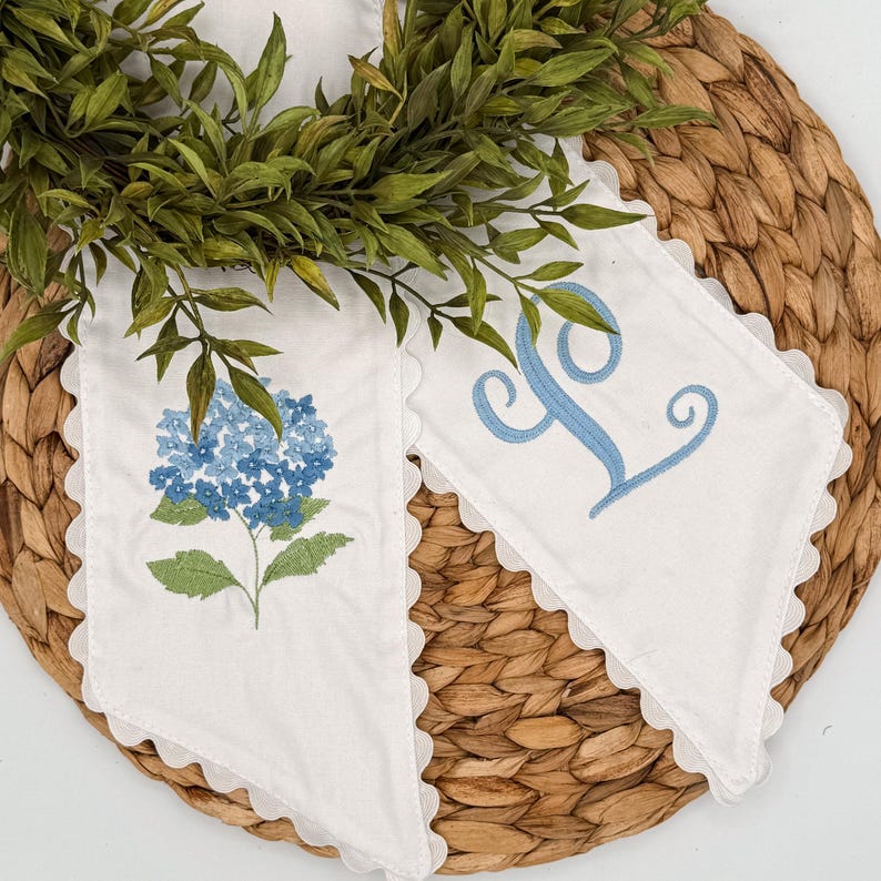May include: Two white decorative fabric pieces with scalloped edges. One features a blue hydrangea embroidery, the other a light blue letter "L". Green foliage and a woven mat form the background.