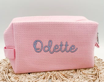 May include: A rectangular light pink cosmetic bag with a waffle-textured fabric surface. The name Odette is embroidered in a cursive, light blue font across the center of the front panel. The bag features a side handle and a zipper closure.