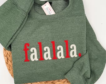 May include: A heathered olive green crewneck sweatshirt featuring the embroidered text "falalala." in alternating red and white letters. The garment includes a small white brand tag attached to the inner neckline with a pink logo design.