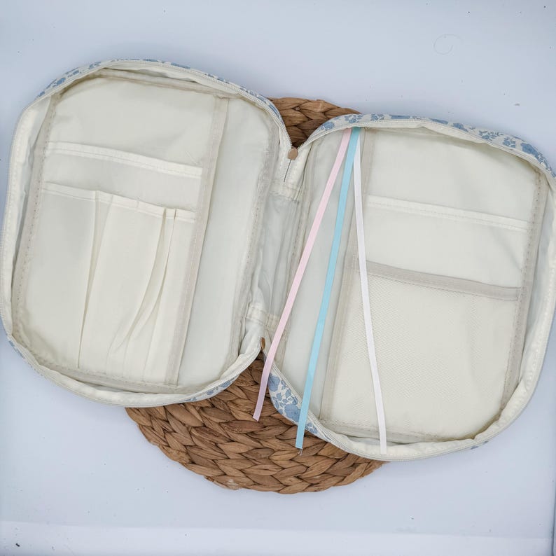 May include: Open, floral-patterned book cover with multiple interior pockets and three colored ribbons. The cover is light blue and white, with a beige interior. The book cover is open, revealing the interior pockets and ribbons.