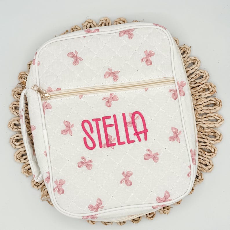May include: White quilted bag with pink bow accents and the name "STELLA" embroidered in pink. The bag has a zipper closure and a handle. The bag is sitting on a woven placemat.