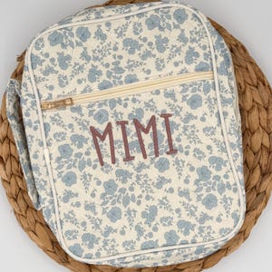 May include: A floral-patterned lunch bag with a cream background and blue flowers. The bag features a gold zipper, a handle, and the name "MIMI" embroidered in a mauve color. The bag is placed on a woven, brown surface.