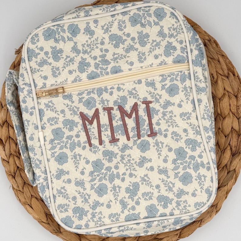 May include: A floral-patterned lunch bag with a cream background and blue flowers. The bag features a gold zipper, a handle, and the name "MIMI" embroidered in a mauve color. The bag is placed on a woven, brown surface.