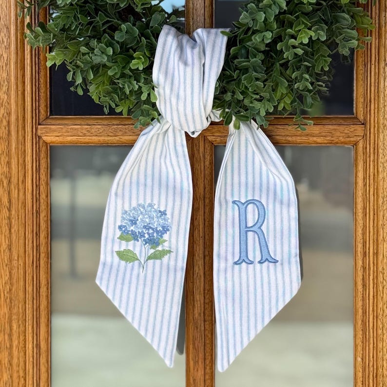 May include: A decorative door hanger featuring a blue and white striped ribbon with a blue hydrangea flower and the letter "R". The ribbon is tied to a green wreath, creating a welcoming aesthetic.