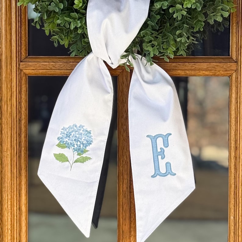 May include: White fabric sash with a blue hydrangea flower and the letter "E" embroidered on it. The sash is tied to a green wreath, creating a decorative accent. The sash is hanging on a wooden door.