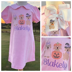 May include: A light pink short-sleeved dress with a scalloped collar featuring an embroidered design of three dogs and a pumpkin, with the name Blakely stitched in purple. A matching white hair bow with a small embroidered dog is also shown.
