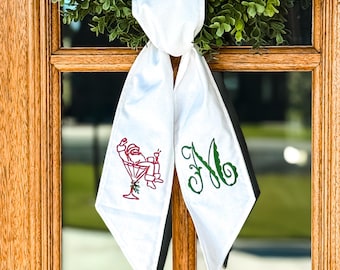 May include: A white fabric ribbon tied in a knot, featuring an embroidered red Santa Claus lounging in a martini glass on the left tail and a large, decorative green embroidered letter M on the right tail, with pointed ends.