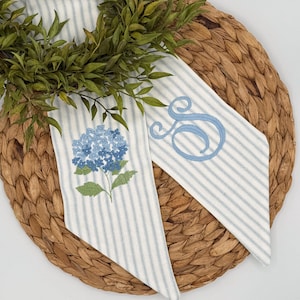 May include: Two decorative fabric pieces with blue and white stripes, one embroidered with a blue hydrangea and the other with a blue script letter "S". The pieces are arranged on a woven, circular placemat with green foliage.