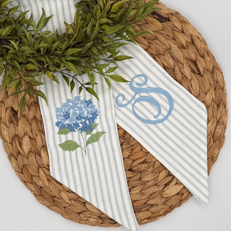 May include: Two decorative fabric pieces with blue and white stripes, one embroidered with a blue hydrangea and the other with a blue script letter "S". The pieces are arranged on a woven, circular placemat with green foliage.