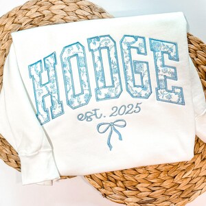 May include: A white sweatshirt featuring the embroidered text HODGE in large block letters filled with a light blue floral pattern. Below the name, the text est. 2025 is embroidered in light blue, accompanied by a small light blue bow graphic.