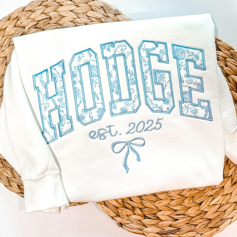 May include: A white sweatshirt featuring the embroidered text HODGE in large block letters filled with a light blue floral pattern. Below the name, the text est. 2025 is embroidered in light blue, accompanied by a small light blue bow graphic.