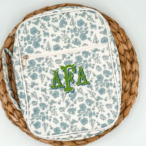 May include: A floral-patterned bag with a white background and blue flowers. The bag has a zipper closure and a handle. The initials "AFA" are embroidered in green with a blue outline. The bag is sitting on a woven mat.