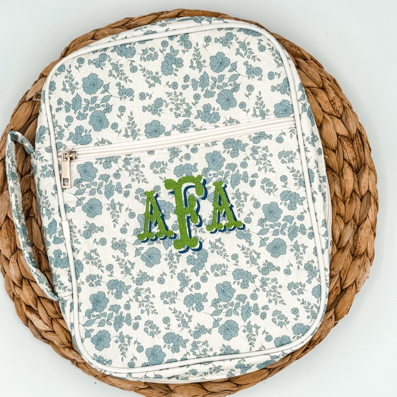May include: A floral-patterned bag with a white background and blue flowers. The bag has a zipper closure and a handle. The initials "AFA" are embroidered in green with a blue outline. The bag is sitting on a woven mat.