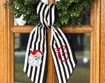 May include: A black and white striped fabric ribbon tied in a knot, featuring an embroidered Santa Claus face on one tail and a red embroidered cursive letter C on the other tail, displayed against a green leafy wreath on a wooden door.
