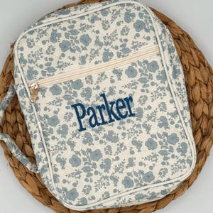 May include: A floral-patterned lunch bag with the name "Parker" embroidered in navy blue. The bag features a cream-colored background with blue flowers, a zipper closure, and a handle. The bag is sitting on a woven mat.