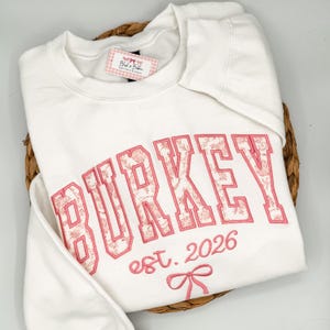 May include: A white crewneck sweatshirt featuring the embroidered name BURKEY in large block letters with a pink floral pattern fill. Below the name, the text est. 2026 is embroidered in pink script, accompanied by a small pink bow graphic.
