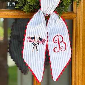 May include: A fabric ribbon with a light blue and white pinstripe pattern and red scalloped edges. One side features an embroidered design of two crossed American flags tied with a black bow, while the other side displays a large red embroidered letter B.