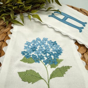 May include: White linen banner with a blue embroidered hydrangea flower and green leaves. A matching banner with a blue embroidered letter "H" is also visible. Both banners have a scalloped edge.