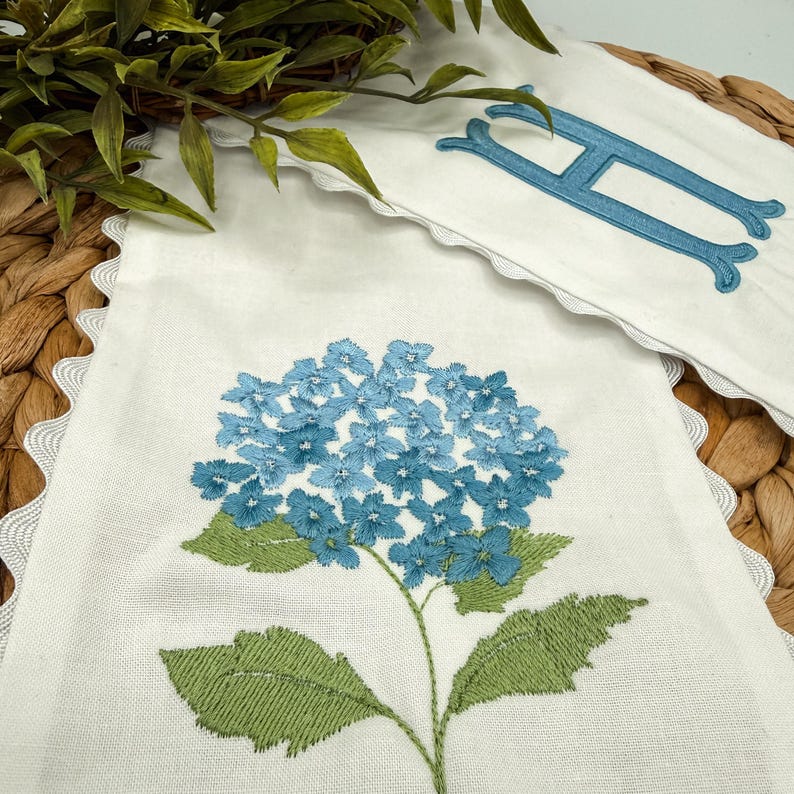 May include: White linen banner with a blue embroidered hydrangea flower and green leaves. A matching banner with a blue embroidered letter "H" is also visible. Both banners have a scalloped edge.