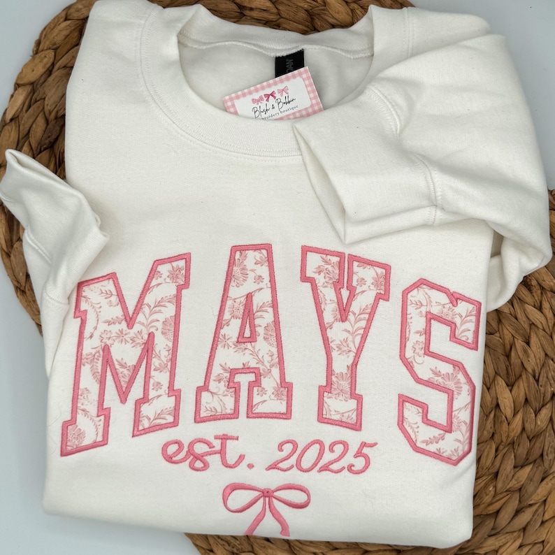 May include: A cream-colored crewneck sweatshirt featuring the word MAYS in large block letters with a pink floral pattern fill and pink outline. Below the text is the phrase est. 2025 and a small pink bow graphic embroidered on the front.