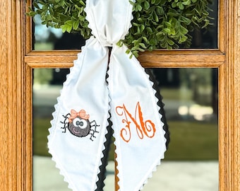 May include: A white fabric door hanger with a scalloped edge featuring an embroidered black spider with a peach bow on the left side and an orange cursive letter M on the right side, tied in a knot at the top and hanging from a green wreath.