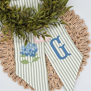May include: Two decorative fabric pieces with green and white stripes, one embroidered with a blue hydrangea and the other with a blue letter "G". The pieces are arranged on a woven placemat and surrounded by a green wreath.