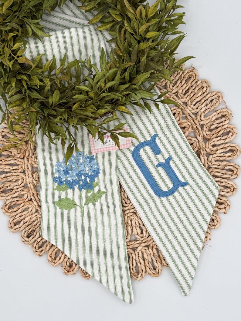 May include: Two decorative fabric pieces with green and white stripes, one embroidered with a blue hydrangea and the other with a blue letter "G". The pieces are arranged on a woven placemat and surrounded by a green wreath.