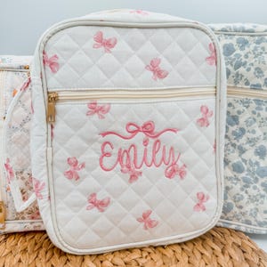 May include: White quilted bag with pink bow accents and the name "Emily" embroidered in pink. The bag has a gold zipper and a rectangular shape. The bag is part of a set with other bags with floral patterns.