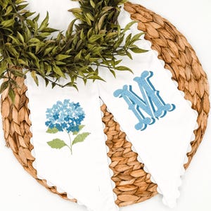 May include: Two white pennant flags with scalloped edges. One flag features a blue hydrangea embroidery, the other a blue monogram letter "M". The flags are displayed on a woven, circular placemat with a green leafy garland.