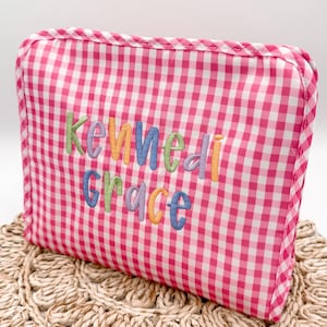 May include: A pink and white gingham cosmetic bag with the name "Kennedy Grace" embroidered in colorful letters. The bag has a rectangular shape with rounded corners and a zipper closure. The bag is sitting on a woven surface.