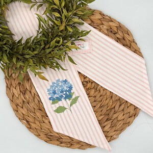 May include: A decorative wreath with a pink and white striped ribbon featuring a blue hydrangea embroidery. The wreath is set on a woven, circular placemat, with a small tag attached to the ribbon.
