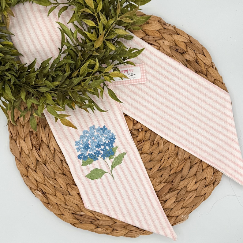 May include: A decorative wreath with a pink and white striped ribbon featuring a blue hydrangea embroidery. The wreath is set on a woven, circular placemat, with a small tag attached to the ribbon.