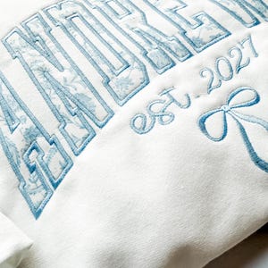 May include: A white sweatshirt featuring large embroidered block lettering filled with a light blue floral pattern. Below the text, the phrase est. 2027 is embroidered in light blue script, accompanied by a light blue embroidered ribbon bow.