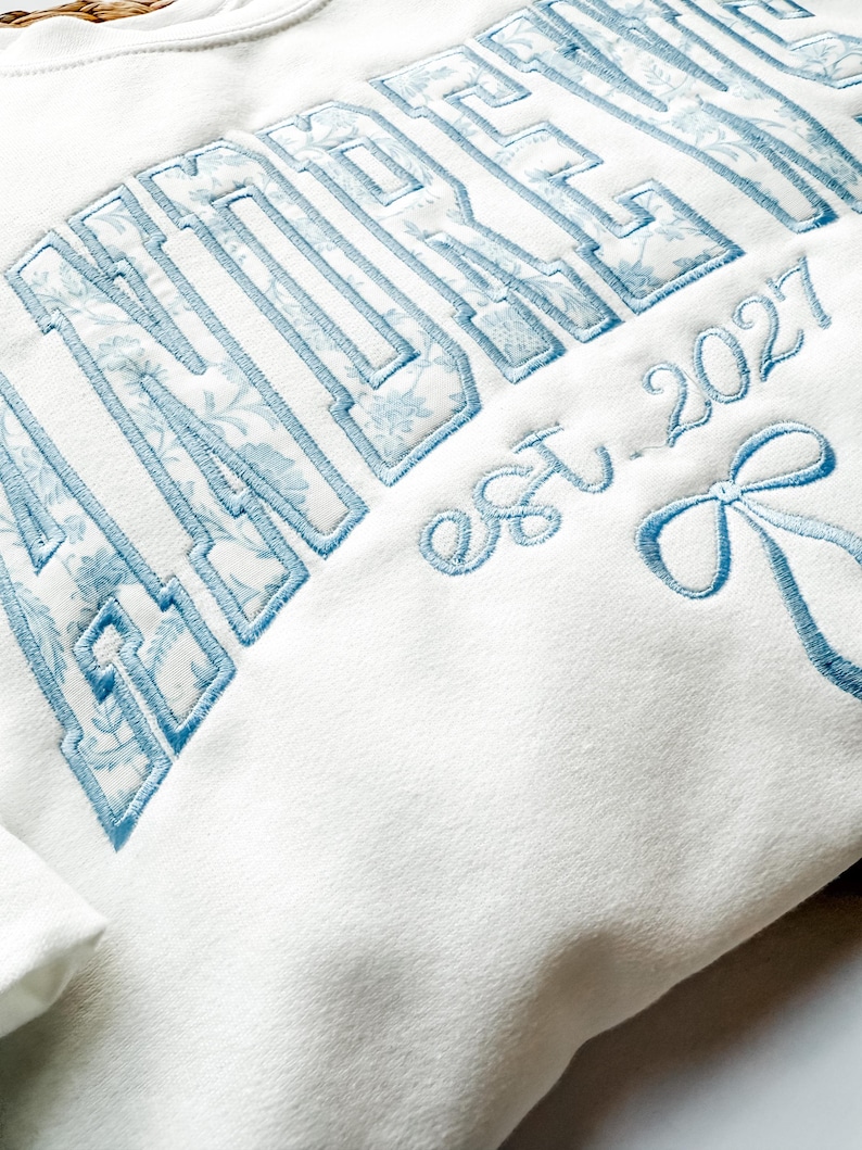 May include: A white sweatshirt featuring large embroidered block lettering filled with a light blue floral pattern. Below the text, the phrase est. 2027 is embroidered in light blue script, accompanied by a light blue embroidered ribbon bow.